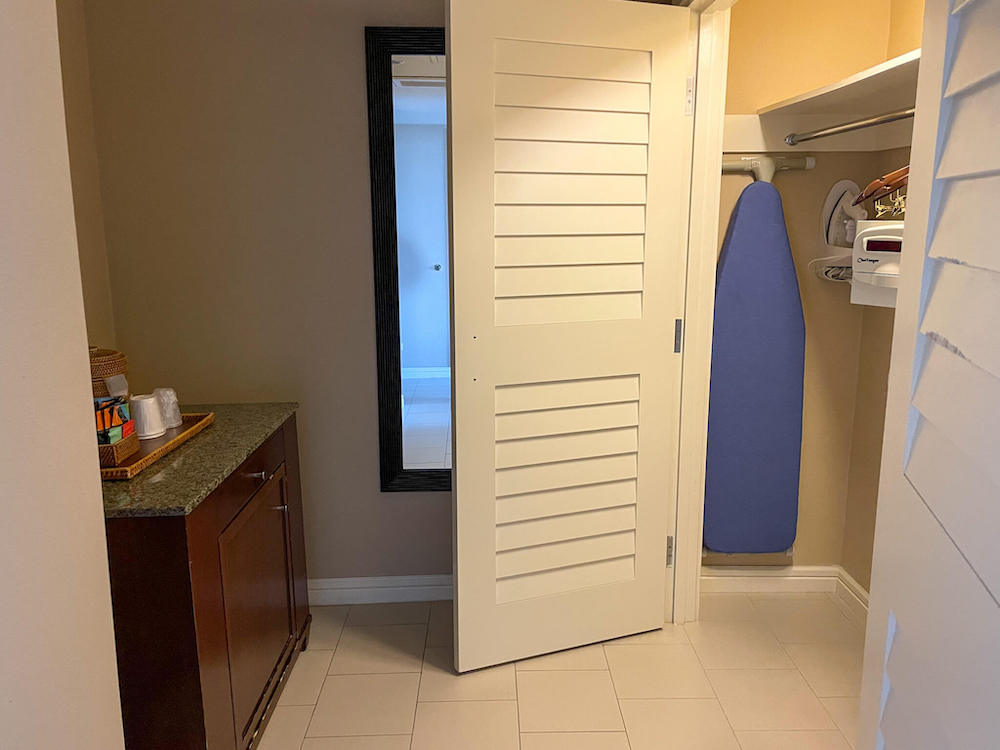 Image of a large closet with an ironing board and safe inside. Plus a coffee maker on a cabinet that has a fridge inside.