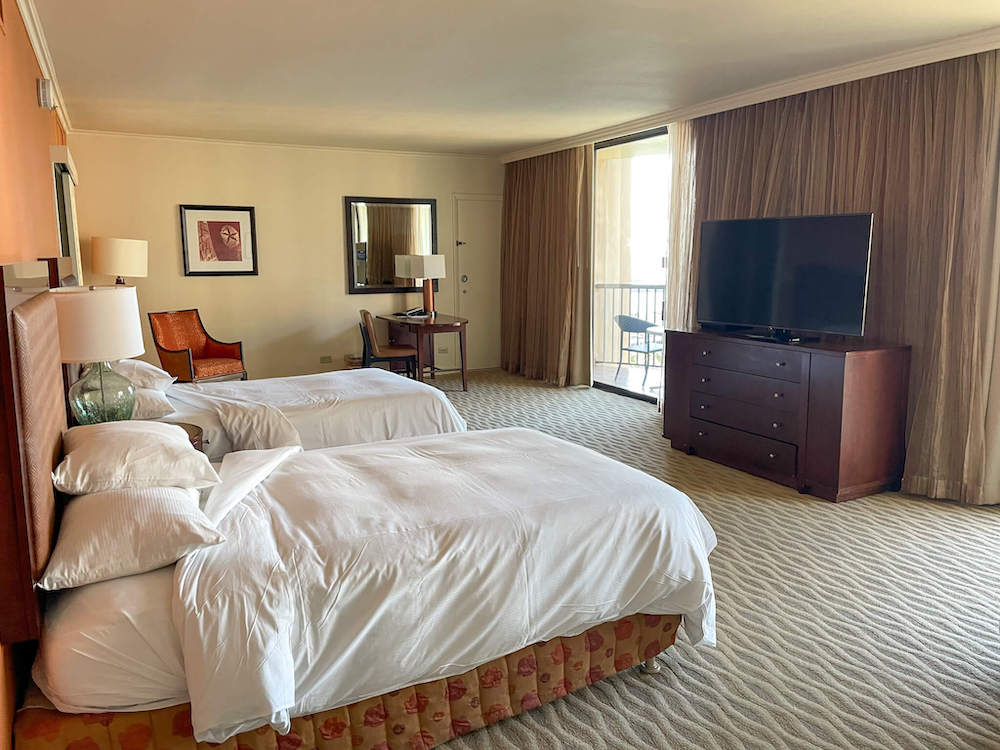 Image of two beds with white linens, a flat screen TV, and some chairs in a hotel room at the Hilton Hawaiian Village.