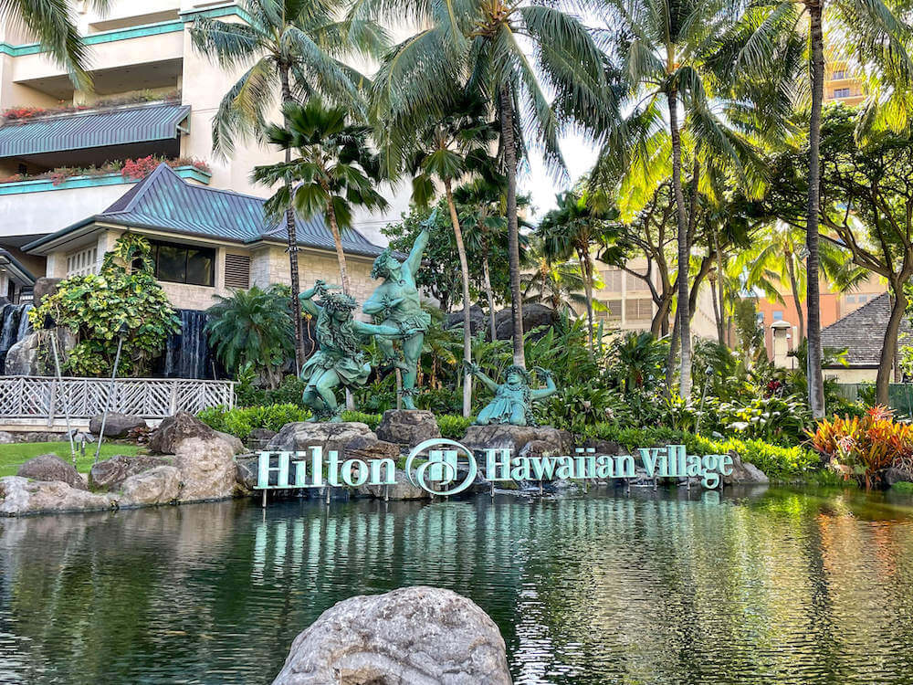 Image of the Hilton Hawaiian Village sign with 3 green statues of hula dancers.