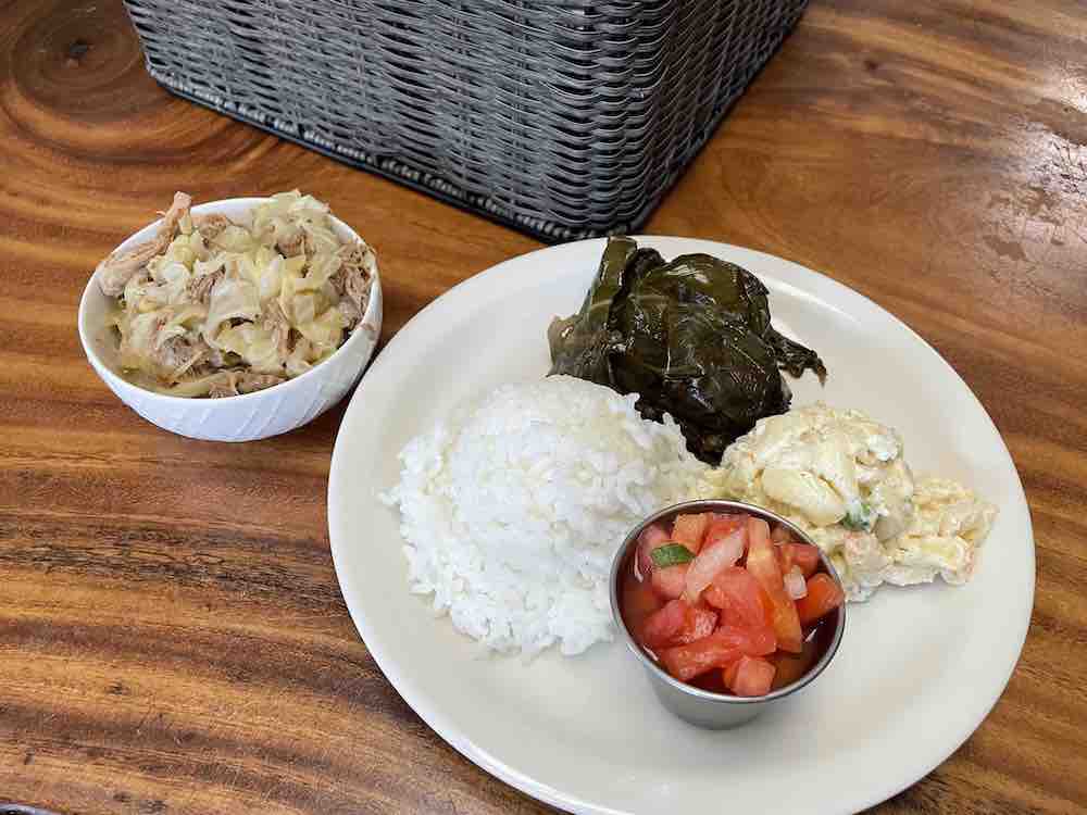 Image of a small bowl of kalua pork and cabbage and a plate with rice, macaroni salad, lomi lomi salmon, and lau lau.