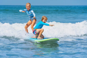 Looking for the best Hawaiian island for kids? Check out this comparison of Maui vs Big Island by top Hawaii blog Hawaii Travel with Kids. Image of two kids on a surfboard in the ocean.