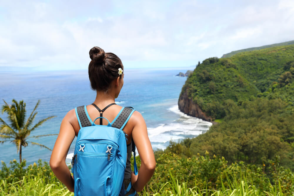 Image of a woman wearing a blue backpack looking out on the Pololu Valley while hiking on the Big Island.