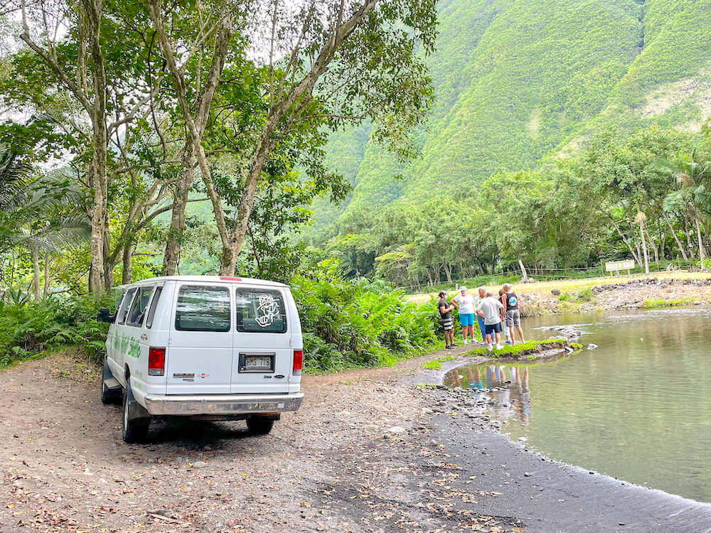 Image of a white van parked along the river with a group of people standing in Waipio Valley on the Big Island of Hawaii.