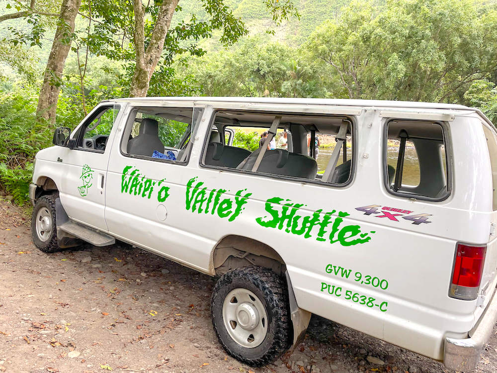 Image of a parked white van with no glass in the windows and the words Waipio Valley Shuttle written on the side in green ink.