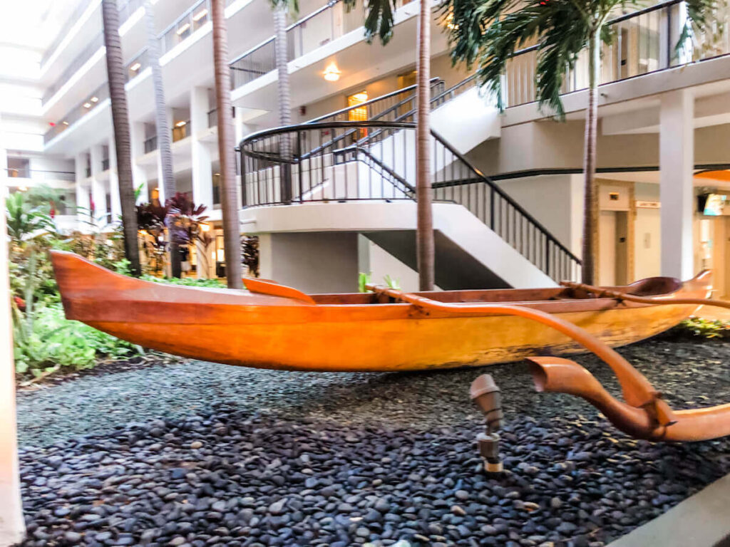 Image of a brown outrigger canoe on a gravel display next to a hotel.