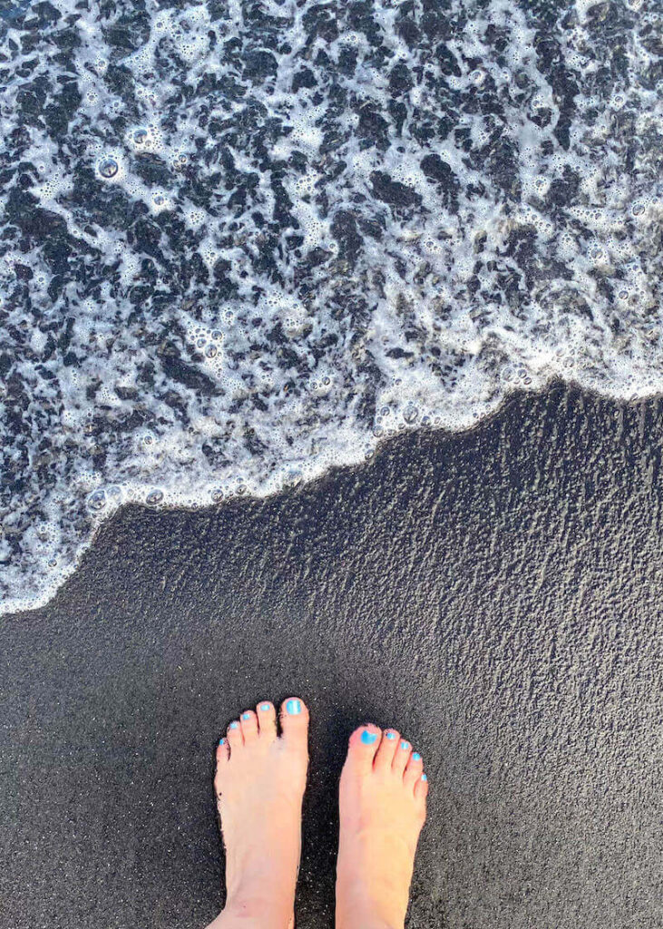 Image of feet with blue toenail polish standing on a Hana black sand beach.