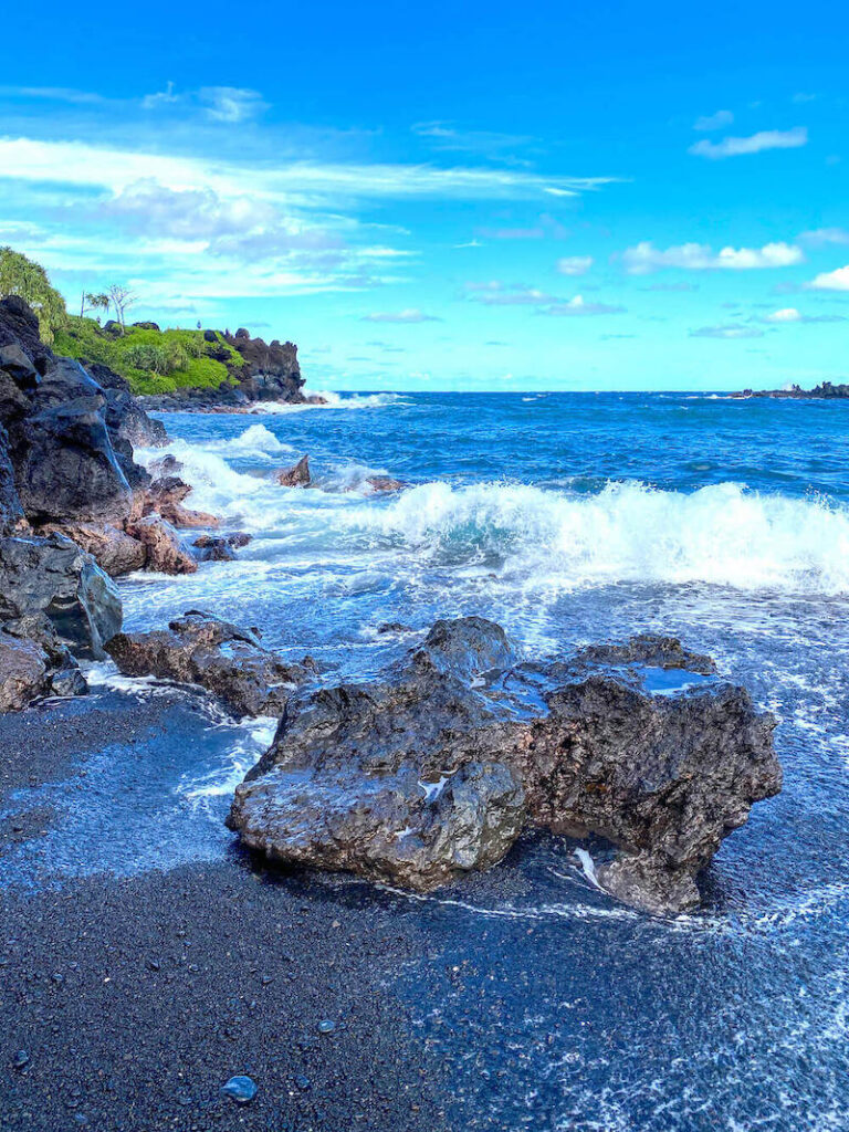 Image of a black sand beach on Maui with waves crashing against the lava rocks.