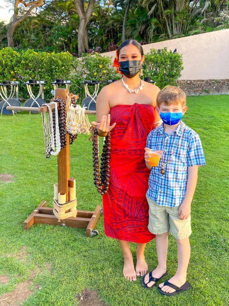 Image of a woman wearing a red Polynesian dress holding kukui nut leis posing with a boy holding a drink at a Maui luau.