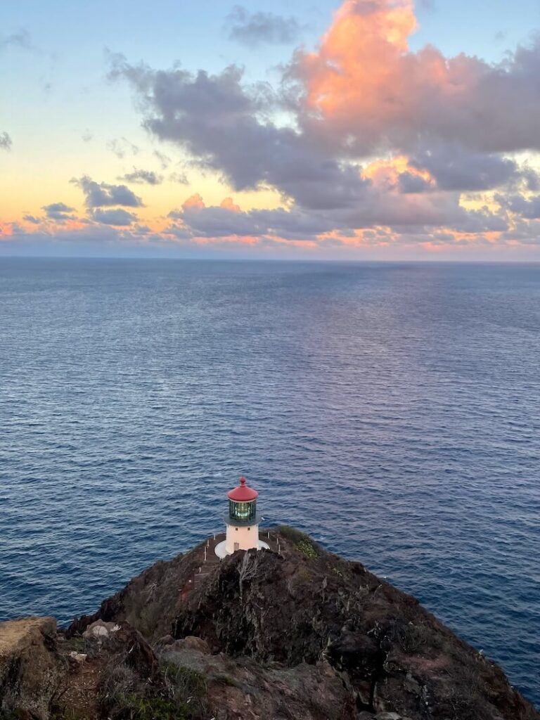 Image of a lighthouse in Oahu with a red roof sitting on a cliff by the ocean.