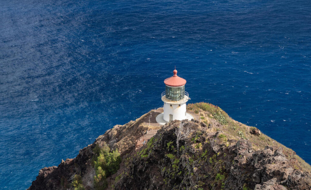 Get tips for doing the Makapuu Lighthouse hike on Oahu with kids by top Hawaii blog Hawaii Travel with Kids. Image of a Hawaii lighthouse with a red roof sitting on a cliff by the ocean.