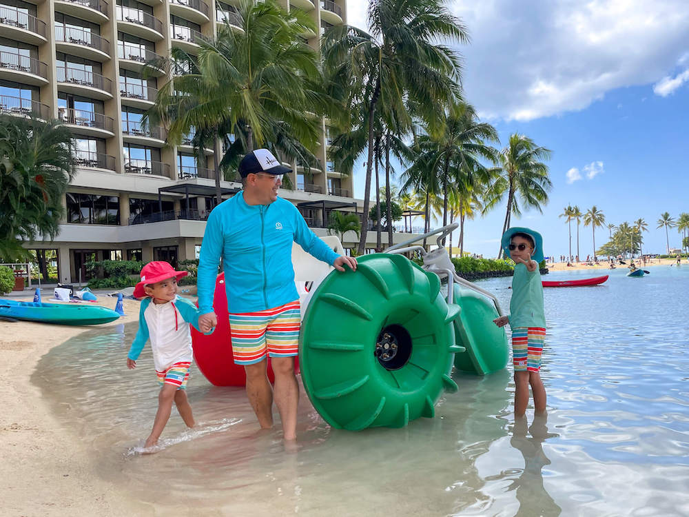 Image of a dad and two boys wearing coordinating rainbow print SPF swimsuits in front of a colorful water trike at the Hilton Hawaiian Village on Oahu.