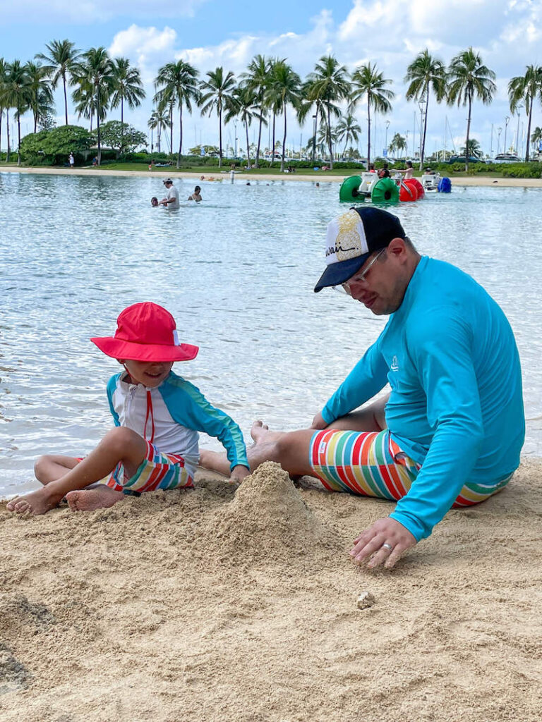 Image of a dad and son wearing coordinating rainbow swimsuits while building a sandcastle in Hawaii.