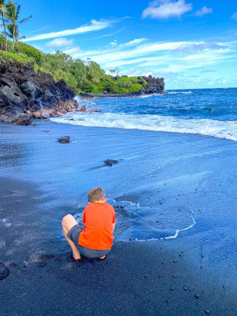 Image of a boy wearing an orange shirt playing at a black sand beach on Maui.