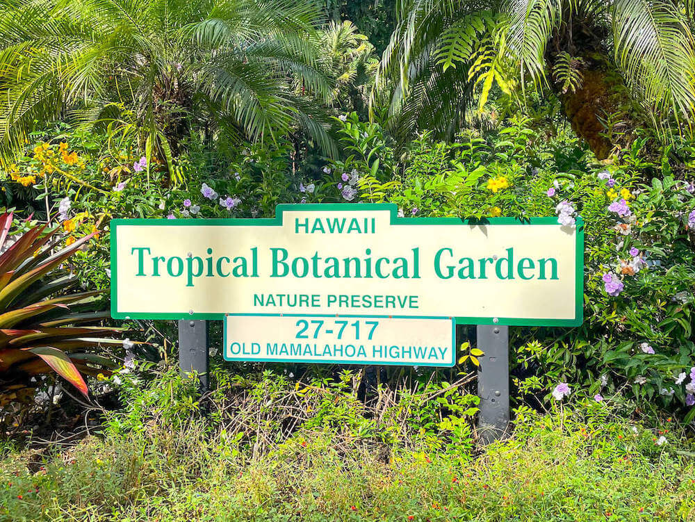 Image of a sign that reads Hawaii Tropical Botanical Garden Nature Preserve 27-717 Old Malamahoa Highway. The sign is surrounded by a tropical garden.
