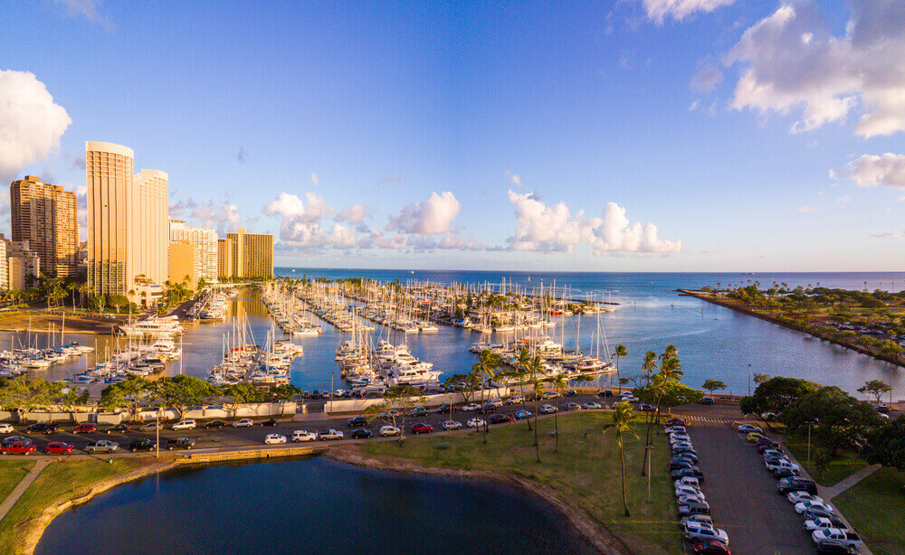 Image of an aerial view of the Ala Wai Boat Harbor and parking lot with Waikiki building and water in the background.