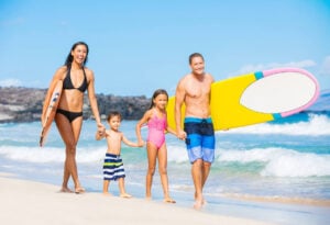Find out the best Hawaiian island for kids recommended by top Hawaii blog Hawaii Travel with Kids. Image of a family with a mom, dad, and two kids holding surfboards and walking along the beach in Hawaii.