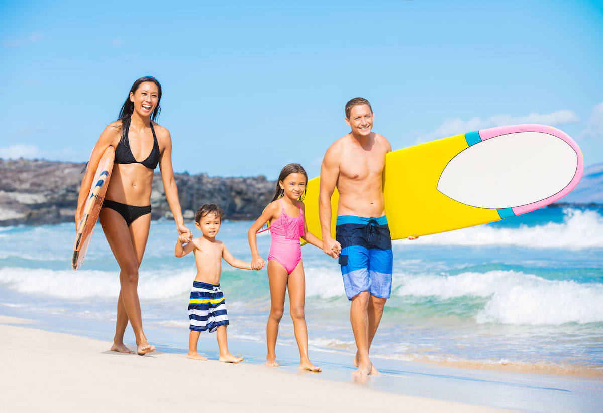 Find out the best Hawaiian island for kids recommended by top Hawaii blog Hawaii Travel with Kids. Image of a family with a mom, dad, and two kids holding surfboards and walking along the beach in Hawaii.