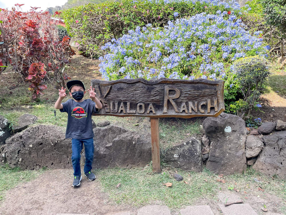 Image of a boy wearing a Jurassic Park shirt in front of a wooden sign that says Kualoa Ranch.