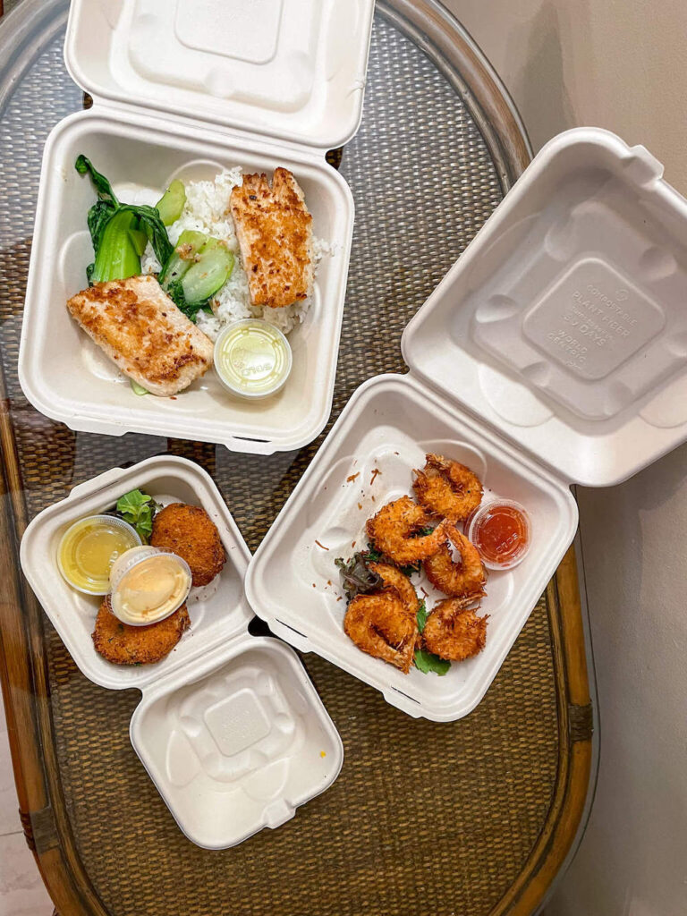 Image of takeout containers on a table with coconut shrimp, crab cakes, and a fish entree from Cafe O Lei in Maui.