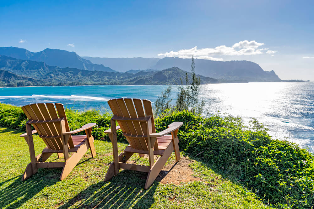 Image of two Adirondack chairs facing the ocean with green tropical mountains in the background.