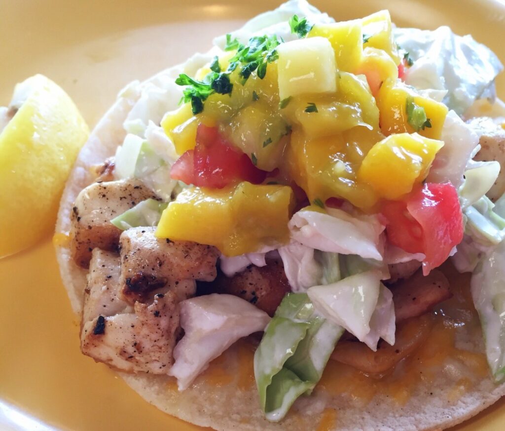 Image of a fish taco topped with coleslaw, tomatoes, and mango.