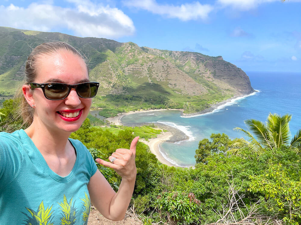 Image of a woman throwing a shaka with an epic view of Halawa Valley in the background.