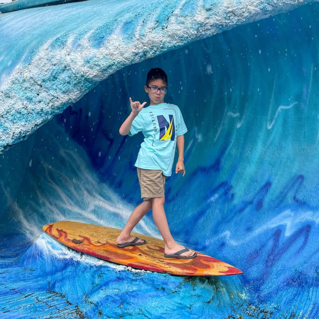 Image of a boy posing for a photo on a surfboard and a fake wave.