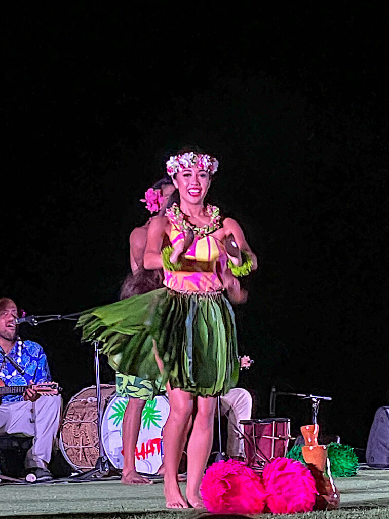 Image of a woman wearing a ti leaf skirt and dancing with hula implements at a Maui luau.