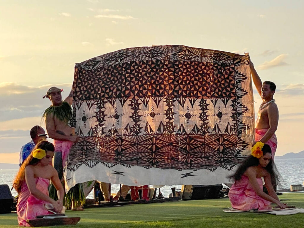 Image of two men holding up a giant piece of Hawaiian barkcloth while two women demonstrate how to beat the cloth.