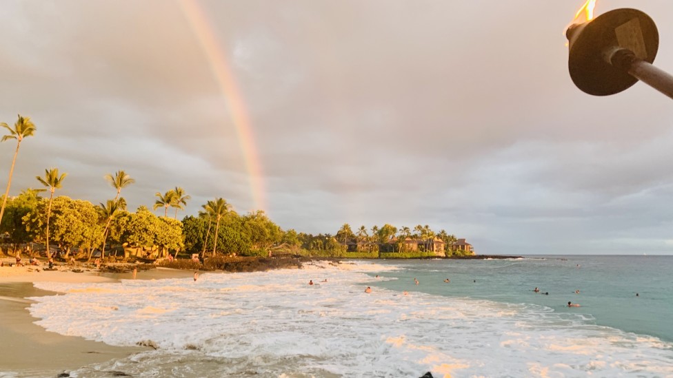 Image of a sandy beach with lots of white ocean foam, a rainbow, and a tiki torch.