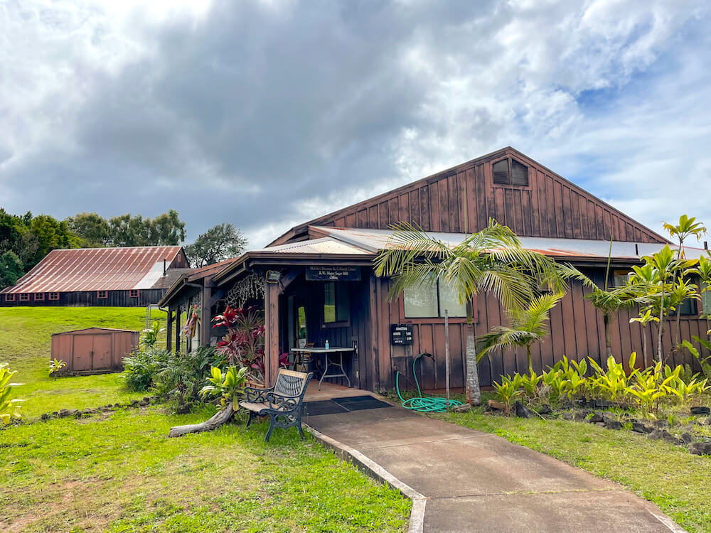 Be sure to visit the Moloka'i Museum on your Maui to Moloka'i day trip. Image of a rustic building in a grassy field with a sugar mill in the background.