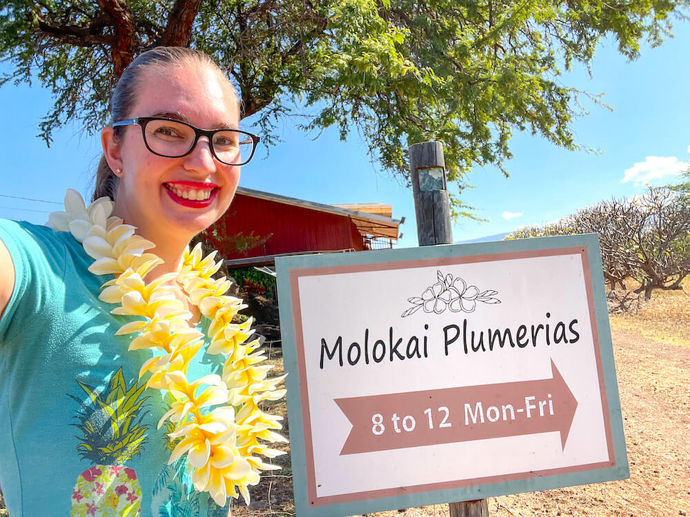 Image of a woman wearing a fresh plumeria lei next to the Molokai Plumerias sign in Hawaii.