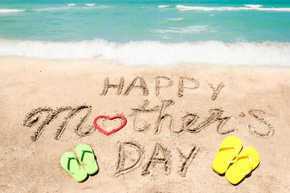 Image of the words Happy Mother's Day written in the sand at a beach with colorful flipflips and a heart shape nearby.