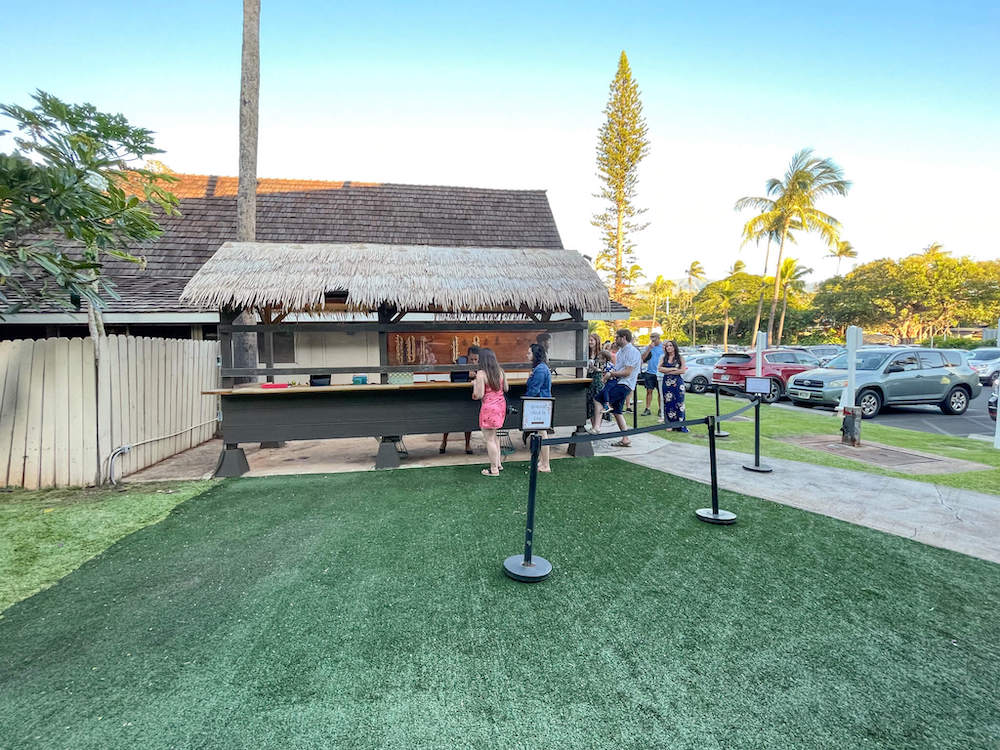 Image of a grass roofed stand with a line of people at the Myths of Maui Luau.