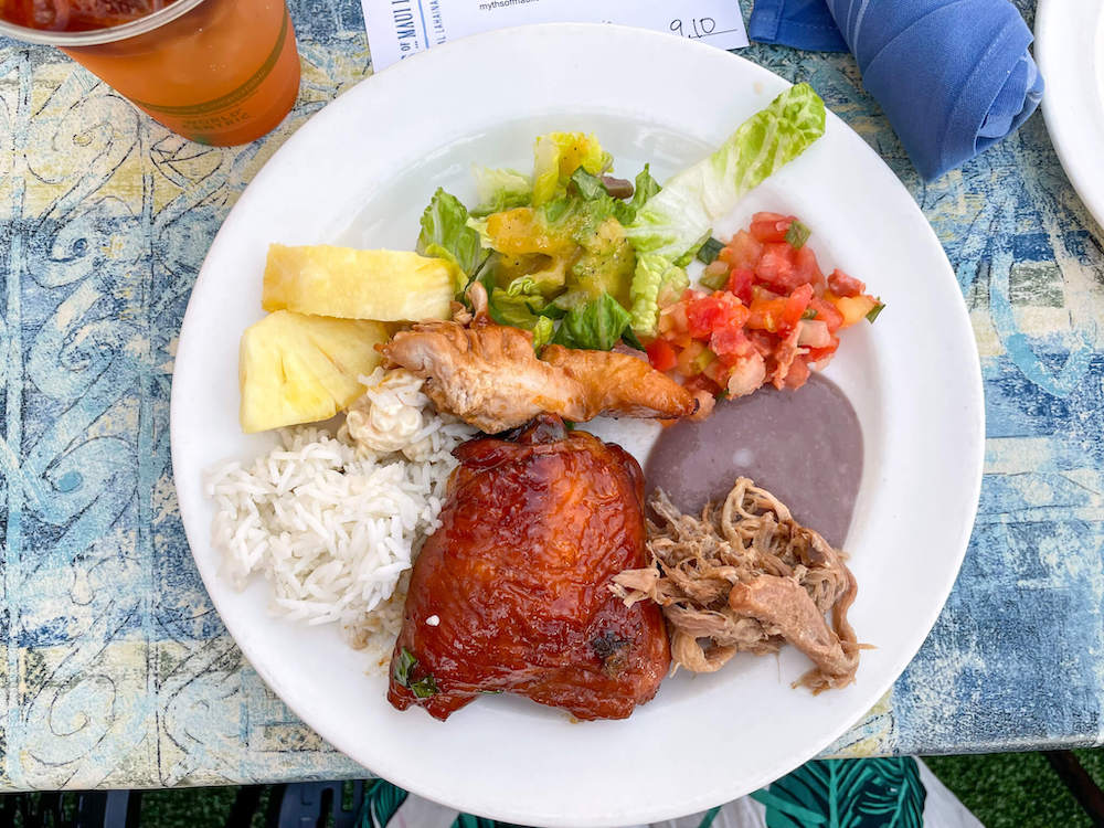 Image of a white plate with pineapple slices, rice, teriyaki chicken, kalua pork, poi, lomi lomi salmon, and salad.