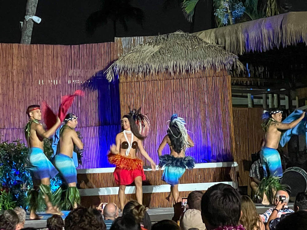 Image of a woman wearing a red costume surrounded by men and women wearing blue costumes as they do Tahitian dancing at the Myths of Maui luau in Lahaina.