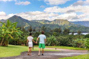 Check out this ultimate guide to North Shore Kauai by top Hawaii blog Hawaii Travel with Kids. Image of a man and woman holding hands while walking on a path in Hanalei Kauai with mountains in background