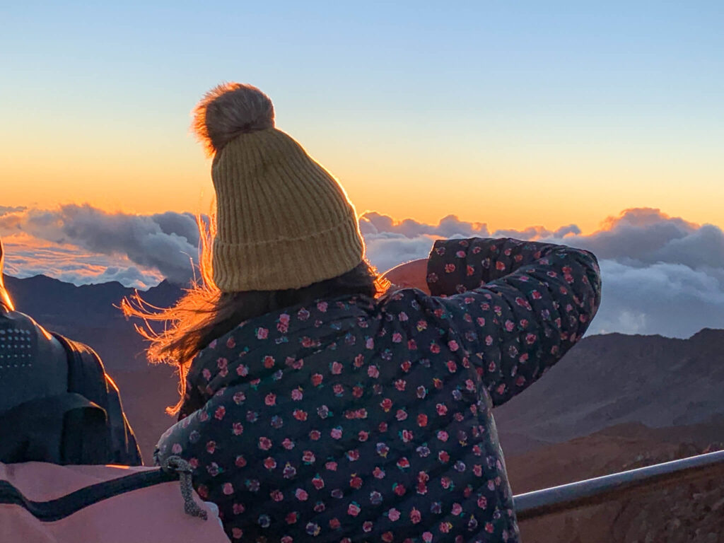 Image of a woman wearing a yellow beanie and a flowery down jacket at sunrise at Haleakala Crater on Maui.