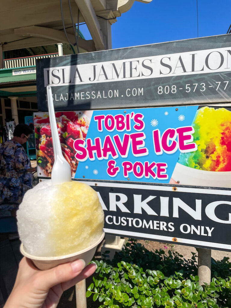 Image of a yellow and white shave ice in front of the Tobi's Shave Ice Sign in Maui.