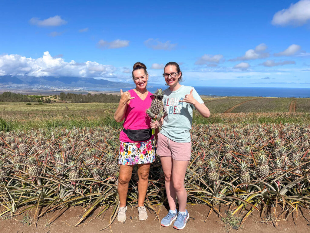 Image of two ladies holding a pineapple and throwing shakas in front of a pineapple field on Maui.