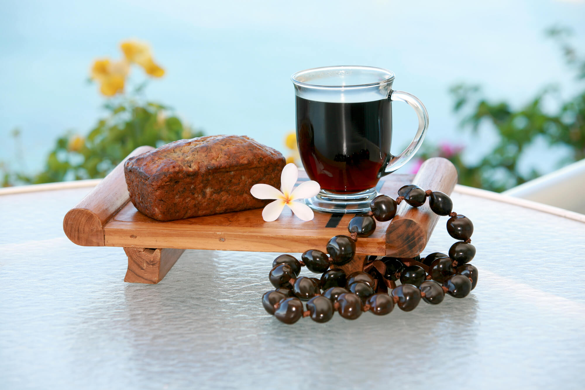 Find out where to eat on Maui with kids recommended by top Hawaii blog Hawaii Travel with Kids! Image of Hawaiian banana bread, coffee, and a kukui nut lei on a table with a tropical background