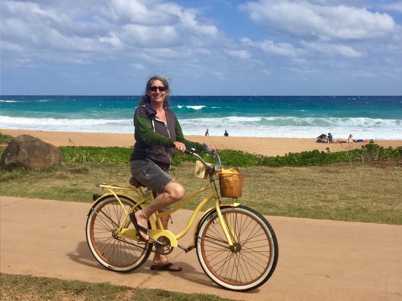 Image of a lady riding a yellow bike on a Kauai bike path in front of the ocean.