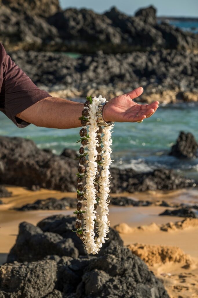 Image of a white flower lei and a kukui nut lei draped on an arm on a beach in Hawaii.