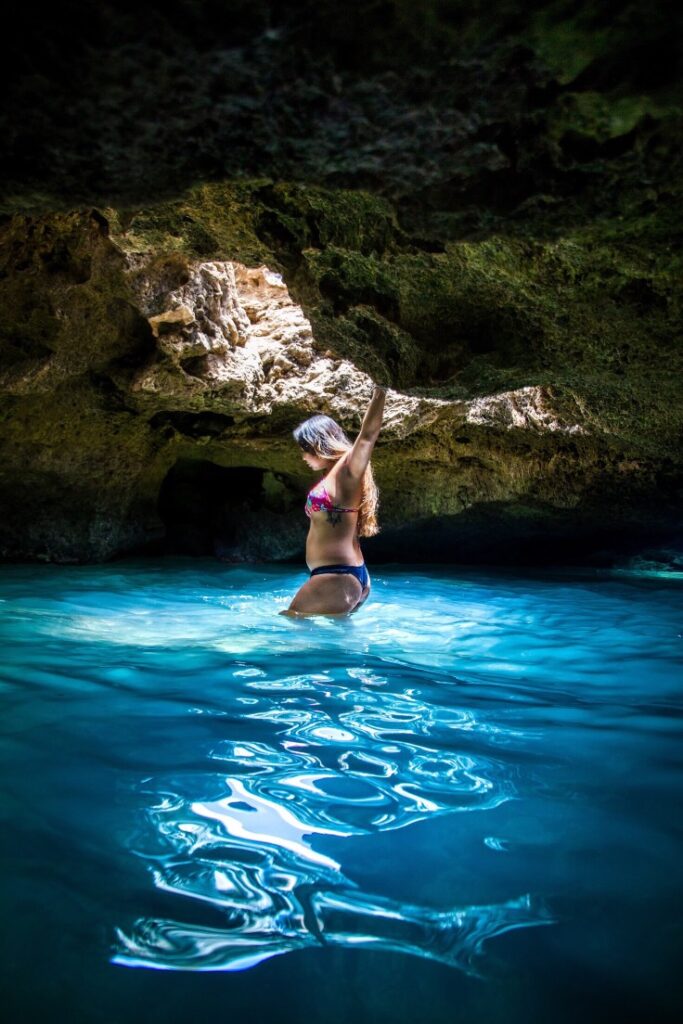 Image of a woman wearing a bikini standing in blue water inside a cave with a skylight.