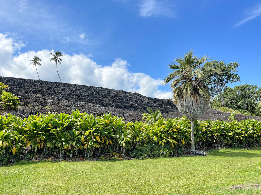 Image of a lava rock temple at a Maui botanical garden with ti leaves and palm trees and grass.