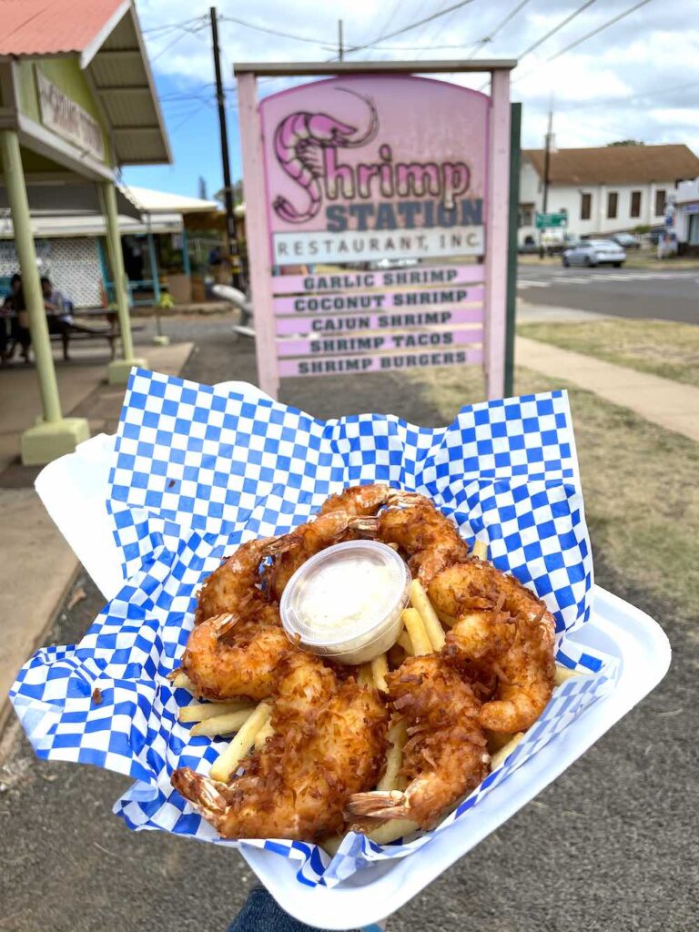 Image of a take-out box with coconut shrimp, french fries, and tartar sauce with the Shrimp Station sign in the background.