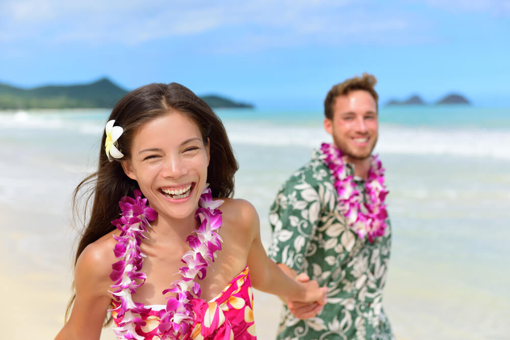 Image of a man and woman smiling while wearing purple orchid leis on a beach in Hawaii.