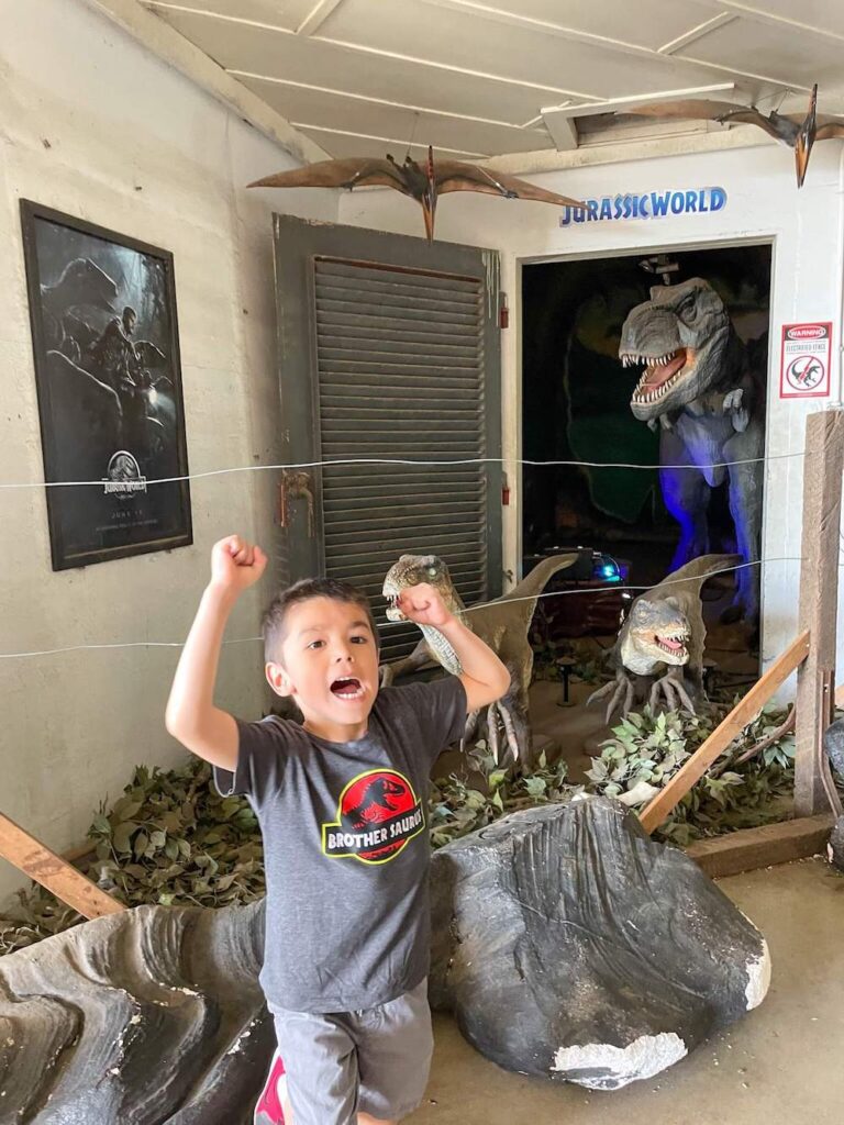 Image of a boy wearing a Jurassic Park shirt in front of a Jurassic World exhibit at Kualoa Ranch.