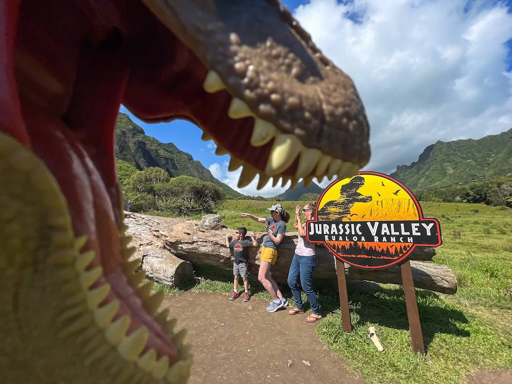 Image of a grandma, mom, and son looking like they are being eaten by a toy dinosaur with the Jurassic Valley Kualoa Ranch sign.