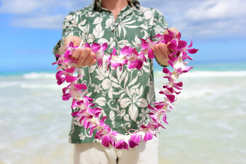 Image of a man wearing a green Aloha shirt holding out a purple orchid lei.